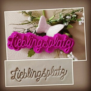 Könnte beinhalten: Ein rosa Plastik-Ausstecher mit den Worten "Lieblingsplatzy" in einer kursivschriftlichen Schriftart. Der Ausstecher befindet sich auf einer weißen Oberfläche mit einem weißen Schmetterling.
