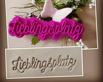 May include: A pink plastic cookie cutter with the words "Lieblingsplatzy" in a cursive font. The cookie cutter is on a white surface with a white butterfly.
