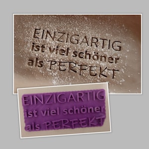 Könnte beinhalten: Ein lila Silikonstempel mit dem Text "EINZIGARTIG ist viel schöner als PERFEKT" in erhabenen Lettern. Der Stempel wird verwendet, um einen Abdruck in Puderzucker zu erstellen.