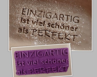 May include: A purple silicone stamp with the text "EINZIGARTIG ist viel schöner als PERFEKT" in raised lettering. The stamp is used to create an impression in powdered sugar.