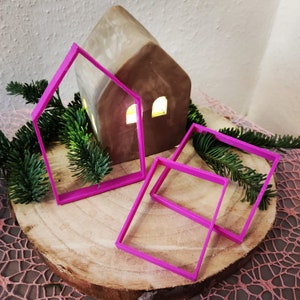 Könnte beinhalten: Drei rosa Plastik-Ausstecher in Form eines Hauses und zweier Quadrate. Die Ausstecher liegen auf einer Holzoberfläche mit einem kleinen, braunen Keramikhaus im Hintergrund.