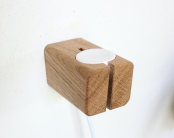 May include: A wooden wall mount for charging an Apple Watch. The mount has a slot for the charging cable and a cutout for the watch face.