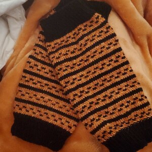 May include: A pair of black and orange striped knitted arm warmers. The arm warmers are folded and lying on a fuzzy orange blanket.