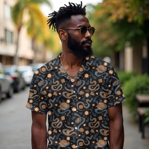 May include: Black button-down shirt with a repeating pattern of gray snakes, dice, and floral elements. The short-sleeved shirt has a camp collar and is worn by a person wearing sunglasses and a necklace.