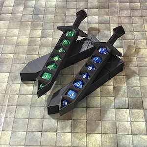 May include: Two black sword-shaped containers for polyhedral dice. Each sword features a series of hexagonal cutouts holding a set of seven dice. One sword contains green dice with white numbers, and the other contains blue dice with white numbers.