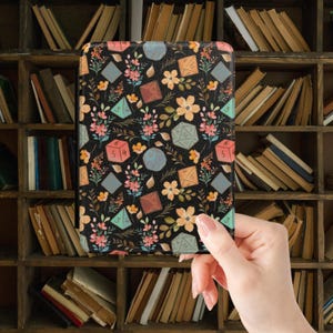 May include: A black e-reader cover with a floral and dice design. The pattern incorporates pink, orange, and yellow flowers, alongside dice shapes in blue, green, and brown. The cover is held in front of a bookshelf.