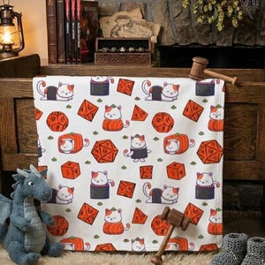 Op de afbeelding: Witte deken met een herhalend patroon van oranje dobbelstenen, sushi en cartoonkatten. De katten hebben verschillende poses en outfits. De deken is gedrapeerd over een houten bank, met een blauw draakspeelgoed op de voorgrond.