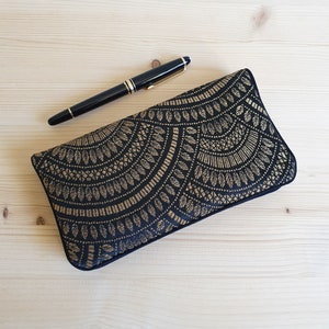 May include: A black clutch with a gold leaf pattern and a black pen with gold accents. The clutch is rectangular and has a decorative design. The pen is black with gold trim and a gold nib. The items are on a light wood surface.