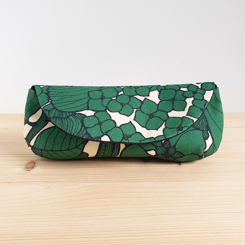 May include: A rectangular fabric case with a rounded flap closure, featuring a repeating pattern of green hydrangea-like flowers and dark green leaves on a cream-colored background. The case has a structured shape and a textured textile finish.
