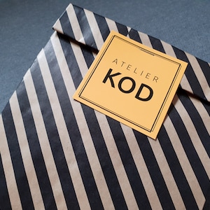 May include: A rectangular paper envelope featuring a diagonal black and tan striped pattern. A square, light orange label is centered on the front, displaying the text ATELIER KOD in bold black capital letters within a thin black border.