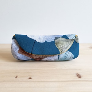 May include: A rectangular fabric pouch featuring a deep teal background with a pattern of large, stylized ginkgo leaves in shades of light blue, white, and brown. The pouch has a curved flap closure with a thin mustard yellow trim along the edge.