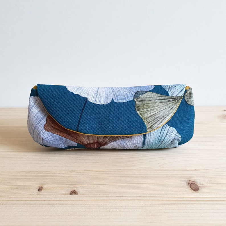 May include: A rectangular fabric pouch featuring a deep teal background with a pattern of large, stylized ginkgo leaves in shades of light blue, white, and brown. The pouch has a curved flap closure with a thin mustard yellow trim along the edge.