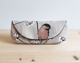 May include: A beige linen eyeglass case with a black and pink bird design. The case has a flap that closes with a magnetic closure.