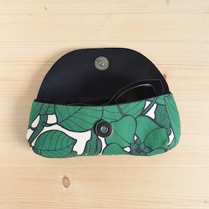 May include: A semi-circular eyeglass case featuring a green and white botanical leaf pattern. The case is open, revealing a black interior lining and a pair of black sunglasses tucked inside. It includes a silver-toned magnetic snap closure.