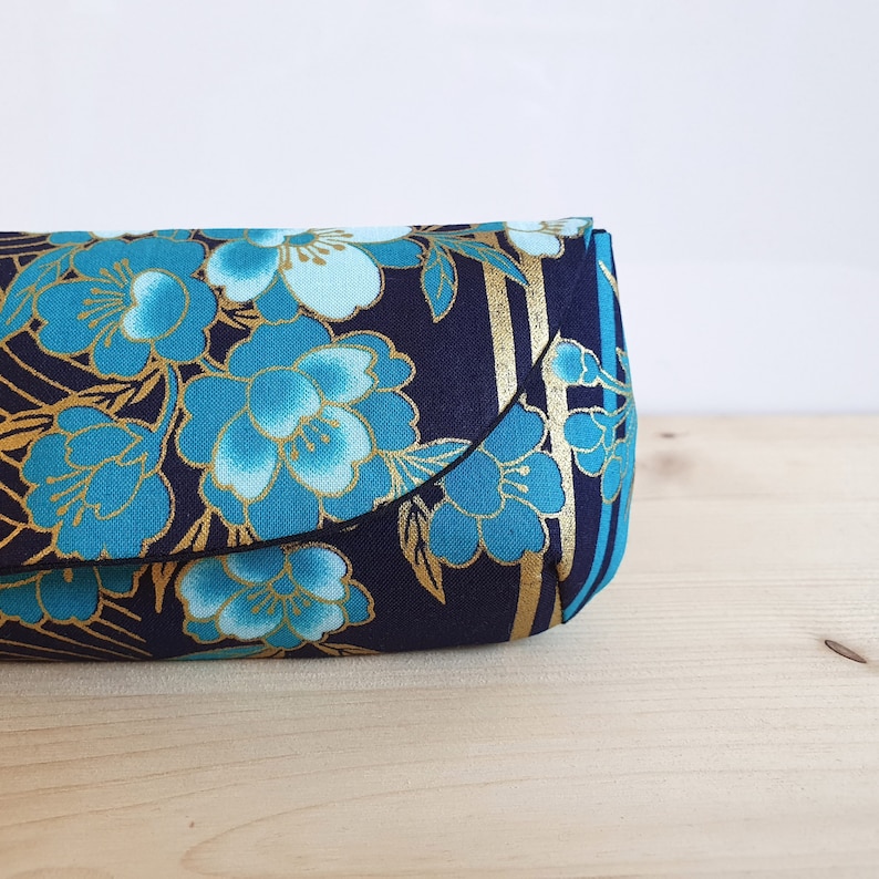 May include: A small, navy blue clutch purse with a gold and blue floral pattern. The purse has a curved shape and a gold zipper.