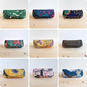 May include: A collection of nine rectangular fabric spectacle cases featuring various patterns and colours, including green floral, teal leaf, navy blue floral, red and green botanical, yellow paisley, black geometric, yellow leaf, floral cat, and blue leaf.