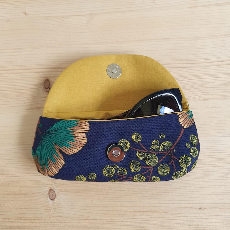 May include: A navy blue fabric glasses case featuring a ginkgo leaf and floral pattern in teal, gold, and yellow. The interior is lined with solid mustard yellow fabric and includes a silver-toned magnetic snap closure on the front flap.