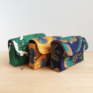 Può includere: Tre piccole pochette rettangolari in tessuto con chiusure a patta. Le pochette presentano colorati motivi botanici in verde, giallo e blu navy. Sono disposte su una superficie di legno chiaro, mettendo in mostra i loro design e le loro trame uniche.