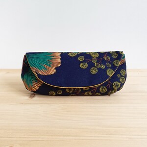 May include: A rectangular fabric pouch with a curved flap closure, featuring a dark navy blue background printed with a pattern of teal and copper-colored ginkgo leaves alongside clusters of small yellow circular floral motifs.
