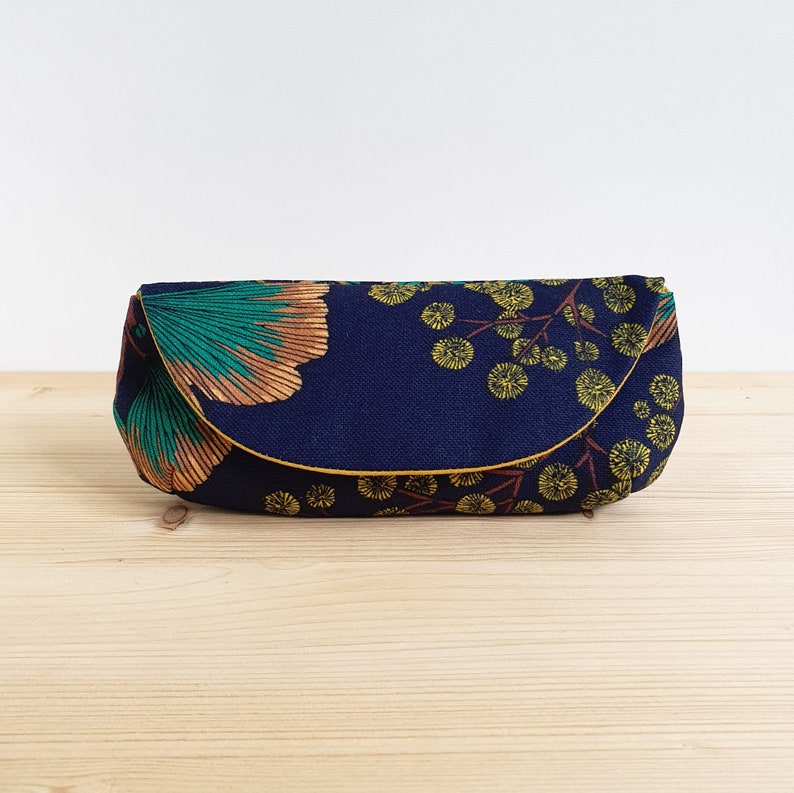 May include: A rectangular fabric pouch with a curved flap closure, featuring a dark navy blue background printed with a pattern of teal and copper-colored ginkgo leaves alongside clusters of small yellow circular floral motifs.