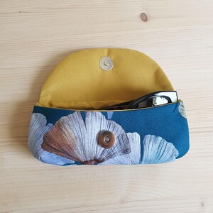 May include: A fabric glasses case with a teal exterior featuring a white and brown ginkgo leaf pattern and a mustard yellow interior lining. The case has a curved flap secured by a silver-toned magnetic snap closure and contains black glasses.