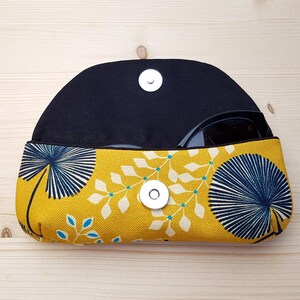 May include: A semi-circular fabric glasses case featuring a mustard yellow background with a dark blue fan-shaped leaf pattern and white foliage accents. The case has a black interior lining and a silver-colored magnetic snap closure at the center.