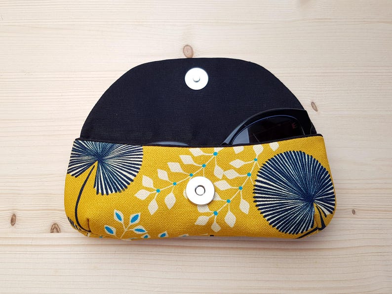 May include: A semi-circular fabric glasses case featuring a mustard yellow background with a dark blue fan-shaped leaf pattern and white foliage accents. The case has a black interior lining and a silver-colored magnetic snap closure at the center.