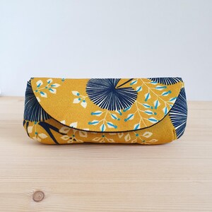 May include: A rectangular fabric pouch with a curved flap closure, featuring a mustard yellow background with a dark navy blue fan-shaped pattern and white leafy branches with small light blue accents throughout the textile design.