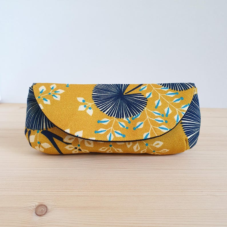 May include: A rectangular fabric pouch with a curved flap closure, featuring a mustard yellow background with a dark navy blue fan-shaped pattern and white leafy branches with small light blue accents throughout the textile design.