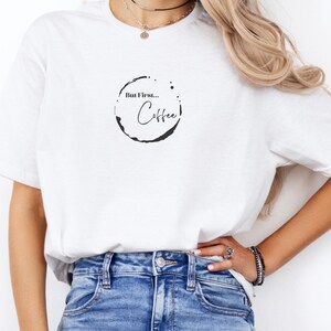 May include: White t-shirt with the phrase "But First... Coffee" in a black, coffee-ring-shaped design. The shirt is a casual, everyday wear item.