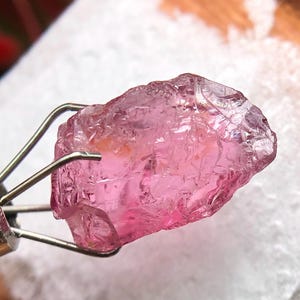 Puede incluir: Una piedra preciosa rosa translúcida en bruto sujeta por una herramienta de metal plateado de cuatro puntas. El cristal presenta una forma irregular y dentada con fracturas internas y facetas visibles, mostrando un tono magenta vibrante.