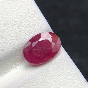 Könnte beinhalten: Ein ovaler, facettierter Edelstein in einer tiefen, gesättigten roten Farbe. Der Stein hat eine polierte Oberfläche mit mehreren geometrischen Facetten, die das Licht reflektieren und seine durchscheinende Qualität sowie den satten Farbton betonen.
