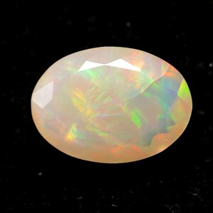 Op de afbeelding: Een ovaalvormige opaal edelsteen met een gefacetteerd oppervlak. De opaal vertoont een melkachtige witte basiskleur met iriserende flitsen van groen, geel en oranje. De edelsteen is geplaatst tegen een zwarte achtergrond.