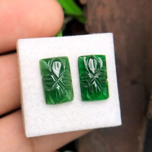 May include: Two rectangular, emerald green jade stones with carved floral designs. The jade pieces are displayed on a white surface. The carvings feature a central flower with symmetrical leaf patterns.