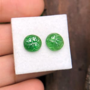 Op de afbeelding: Twee ronde, heldergroene edelstenen met gebeeldhouwde klaverbladmotieven. De edelstenen worden gepresenteerd op een wit oppervlak, waarbij hun levendige kleur en gedetailleerde ontwerpen worden benadrukt. Voor oorbellen of sieraden.
