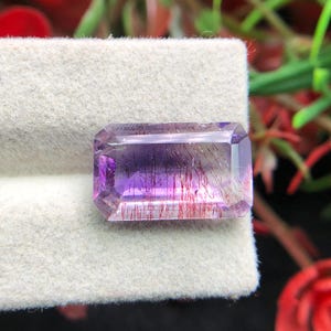 Puede incluir: Una gema rectangular, predominantemente púrpura con inclusiones rojas, sobre un fondo neutro. El cuerpo transparente de la gema permite el paso de la luz, mostrando los intrincados patrones en su interior. El corte de la piedra es preciso.