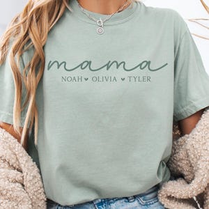 May include: A light green t-shirt with the word "mama" in cursive script, with the names Noah, Olivia, and Tyler below. The shirt is a relaxed fit and has short sleeves. The shirt is made of a soft material.