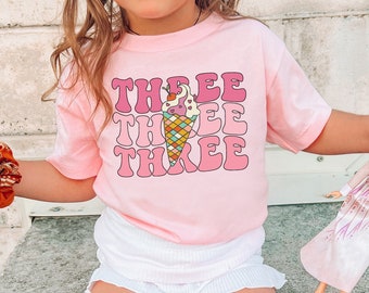 May include: A young child wearing a pink t-shirt with the text "THREE" repeated three times in a retro font. The shirt features a colourful ice cream cone graphic. The child is holding a doll and wearing white socks and trainers.