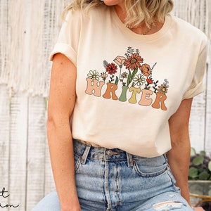 May include: A cream colored t-shirt with a floral design and the word "WRITER" in a retro font.