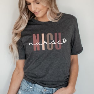 Op de afbeelding: Een donkergrijs T-shirt met de tekst "NICU nurse" in witte letters. De letters "NICU" zijn in een kleurrijk bloklettertype met roze, perzikkleurige en bruine kleuren. Naast het woord "nurse" staan twee kleine witte voetafdrukken.
