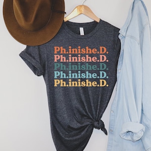 May include: A dark gray t-shirt with the text "Ph.inished.D." repeated five times in a stacked design. The text is in a retro font and is printed in shades of orange, pink, blue, and green.