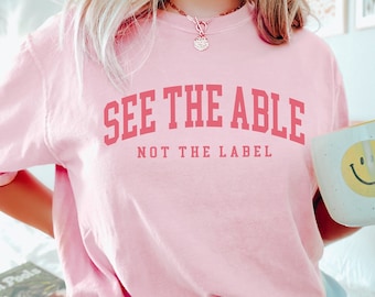 May include: Pink t-shirt with the text "See the Able, Not the Label" in pink lettering. The person is wearing blue shorts and holding a white mug with a yellow smiley face.