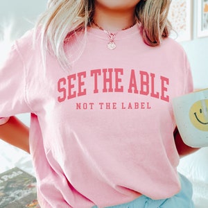 May include: Pink t-shirt with the text "See the Able, Not the Label" in pink lettering. The person is wearing blue shorts and holding a white mug with a yellow smiley face.