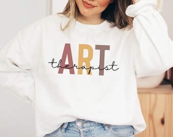 Op de afbeelding: Witte trui met de tekst "ART therapist" in kleurrijke blokletters. De letters zijn in tinten roze, geel en bruin.