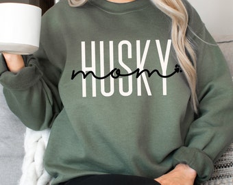 May include: A green sweatshirt with the text "Husky Mom" in white letters with a black paw print. The sweatshirt is being worn by a person sitting on a couch.