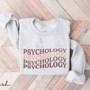 May include: A gray sweatshirt with the word "PSYCHOLOGY" repeated four times in a stacked design. The text is in a retro font and is printed in shades of brown, pink, and peach.