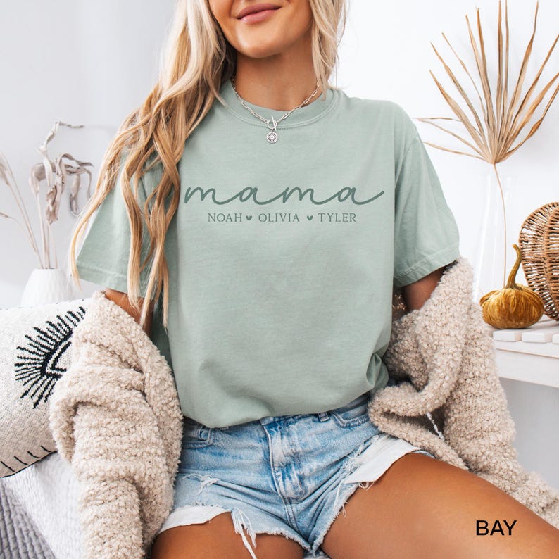 May include: A light green t-shirt with the word "mama" in cursive script, with the names Noah, Olivia, and Tyler below. The shirt is a relaxed fit and has short sleeves. The shirt is made of a soft material.