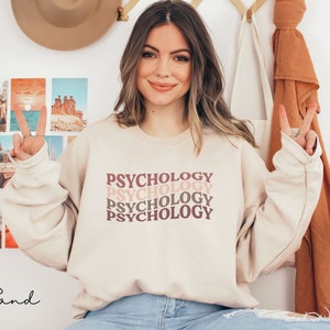 May include: A beige sweatshirt with the word "Psychology" repeated four times in a retro font. The text is in shades of pink and brown.
