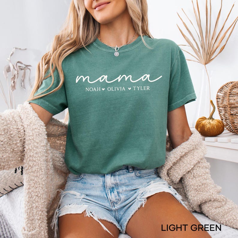 May include: Light green t-shirt with the word "mama" in white script, followed by the names Noah, Olivia, and Tyler. The shirt is a crew neck and is worn with denim shorts. The words "LIGHT GREEN" are at the bottom of the image.