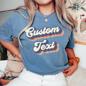 May include: A blue jean t-shirt with a retro-style graphic that says "Custom Text" in rainbow colors.